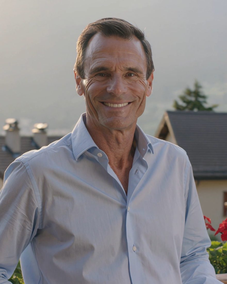 Lächelnder Mann im hellblauen Hemd auf einem Balkon mit Geranien, im Hintergrund Häuser und Berge